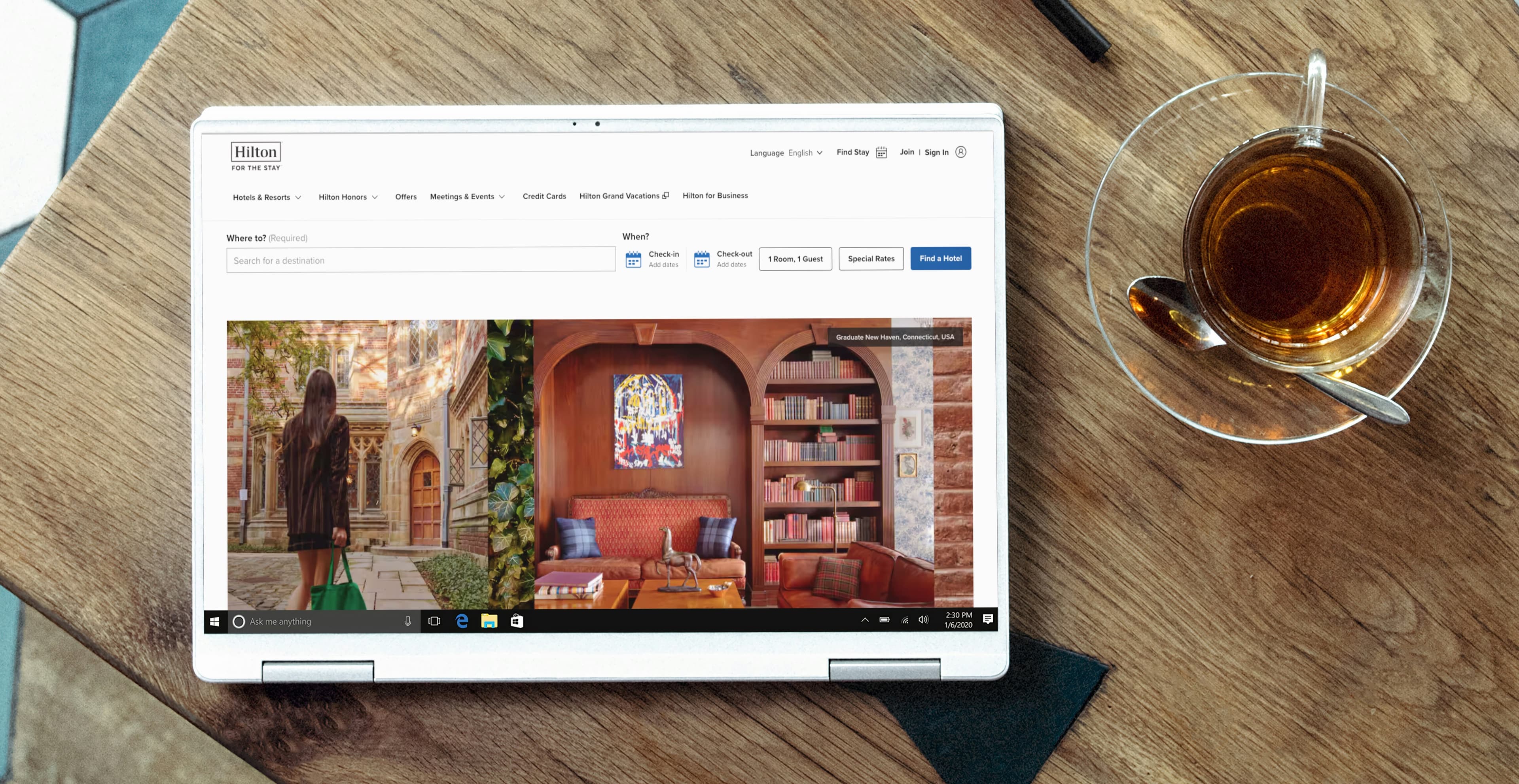 A laptop on a wooden table displays the Hilton website, showing a search bar and calendar icons for booking rooms, and images of a hotel exterior and cozy interior.